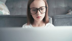 A close-up shot of a serious little 8 years old girl in glasses looking to a laptop screen and typing. Remote education technologies and homework.