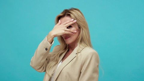 Closeup portrait of nosy curious adult lady with blond hair closing eyes with hand and spying through fingers, hiding and peeping. Indoor studio shot isolated on blue background