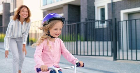 Caucasian beautiful mother teaching small cute girl in helmet riding on bike at street in suburb. Little kid riding the bicycle and learning. Schoolgirl with mommy. Daughter and mom. Parenting.