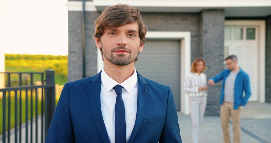 Portrait Caucasian handsome male real-estate agent in suit and tie standing outdoors at big house, smiling and using tablet device. Couple of buyers on background. man tapping and scrolling on gadget. Royalty-Free Stock Footage #1058653282