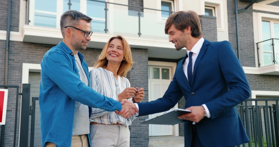 Caucasian male real-estate agent standing outdoors at big house in yard and passing keys to young joyful couple. Male and female buyers of mortgage getting key from new home at suburb. Selling estate. Royalty-Free Stock Footage #1058653285