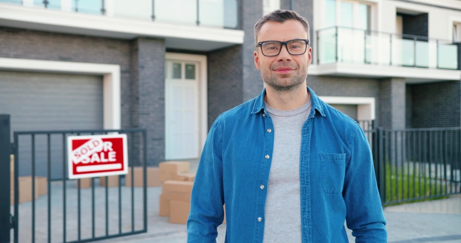Portrait of joyful happy Caucasian man smiling to camera and showing key to camera while moving in new home. Outdoors. Male demonstrating keys. Cardboards on background. Giving thumb up.