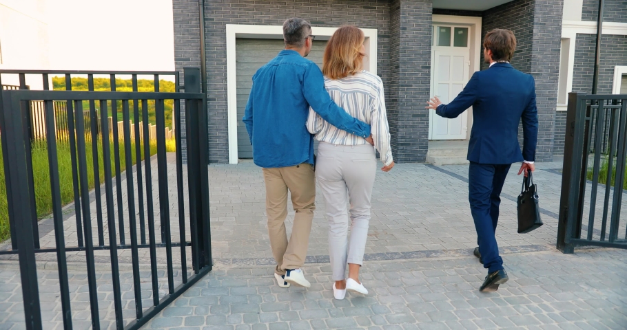 Rear on Caucasian male cheerful real-estate agent in suit and tie walking in yard of big house and showing it to happy just-married couple in hugs. Back view on man and woman observing new home. Royalty-Free Stock Footage #1058653342