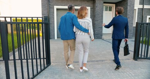 Rear on Caucasian male cheerful real-estate agent in suit and tie walking in yard of big house and showing it to happy just-married couple in hugs. Back view on man and woman observing new home.