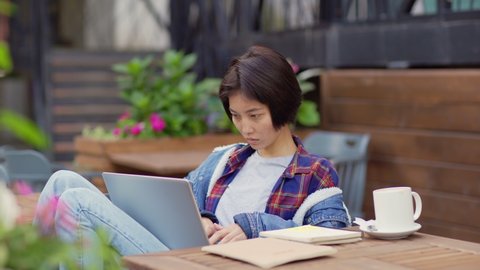 Medium side view shot of young Asian woman working or studying on laptop computer and looking away thoughtfully sitting at table at outdoor cafe