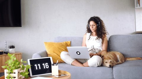 Woman with pet dog sitting indoors at home, using headphones and laptop.