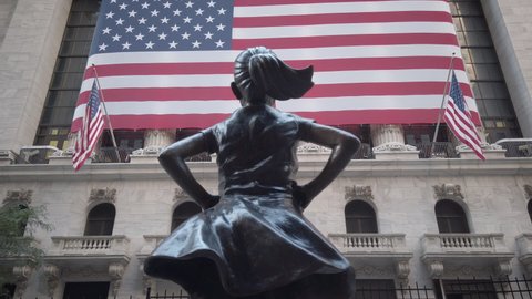 New York / USA - Sept 5, 2020: Fearless Girl statue by Kristen Visbal looking at the New York Stock Exchange (NYSE) with US flags.  Filmed during the Coronavirus pandemic (Covid-19). Empty streets.