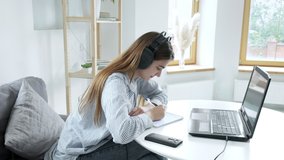 Serious female student wears headphones, studies with internet teacher, learns language. focused young woman talks to tutor, looks at laptop, writes notes. online education, video call tutoring