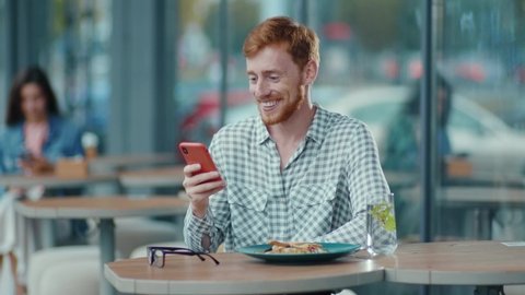 Outdoor scene of positive ginger young man browsing smartphone social network app smiling receiving warm text messages from girlfriend outside in the cafe.