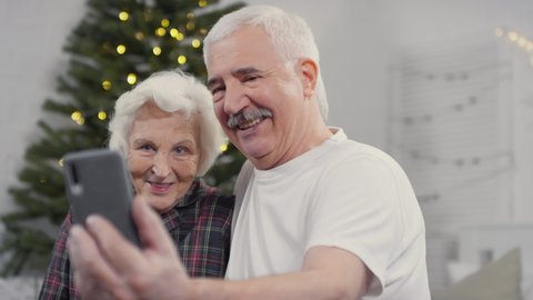 Handheld shot of happy elderly man and woman in sleepwear holding mobile phone and taking selfie or talking on video call with family on Christmas morning