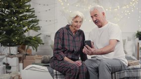 Handheld shot of happy senior man and woman in sleepwear sitting on bed on Christmas morning and video calling their family on mobile phone