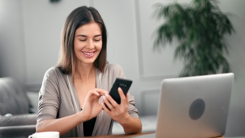 Joyful young woman chatting use smartphone enjoying break during work in front of laptop. Smiling female typing message or texting on mobile sitting on desk at home office. Medium shot on RED camera