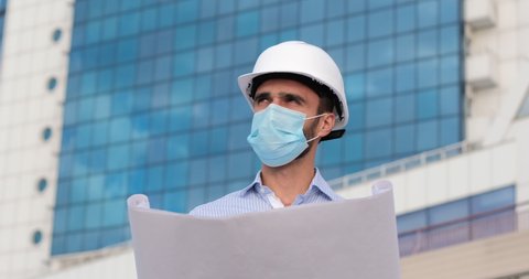 A man in a white protective helmet and face mask unfolds a paper plan, looks at the drawing, and then into the distance at the construction site. A portrait from the lower plan.