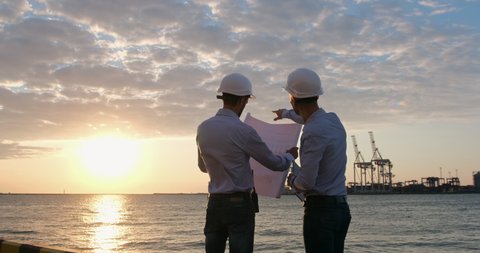 Engineers in white safety helmets discuss a construction plan with cranes in the background at dawn. The man points with his hand to the drawing and then into the distance.