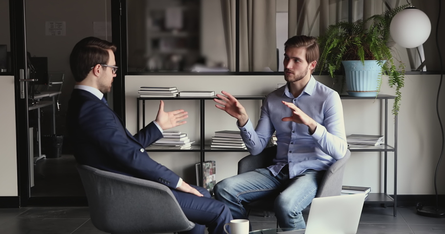 Young confident businessman discussing investment opportunity with financial expert in formal wear, seated on comfortable chairs in office. Smart startup owner consulting with lawyer at meeting.