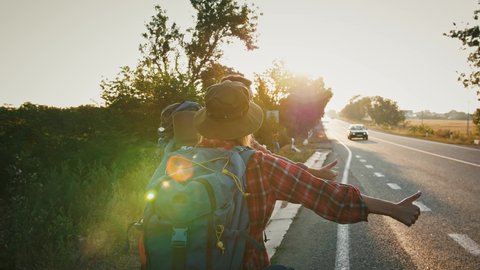 Tracking shot of couple of tourists hitchhiking on highway in evening