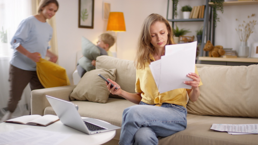 Medium shot of frustrated young woman looking at documents and mobile phone while working from home during pandemic. Playful children running around and pillow fighting Royalty-Free Stock Footage #1058686312