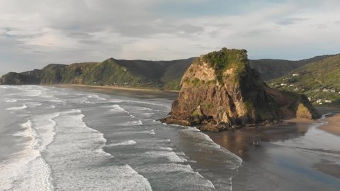 SLOWMO - Breathtaking aerial drone shot of Lion Rock on Piha Beach with surfing waves during sunset