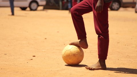 Medium close-up of young boy standing with his bare foot on soccer ball in dusty rural field of dirt before stepping back and kicking it on bright sunny day
