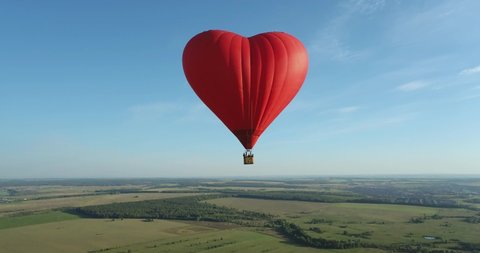 Red hot air balloon in the shape of a heart flight above flat field and forest landscapes at summer sunny day - Aerial drone wide view