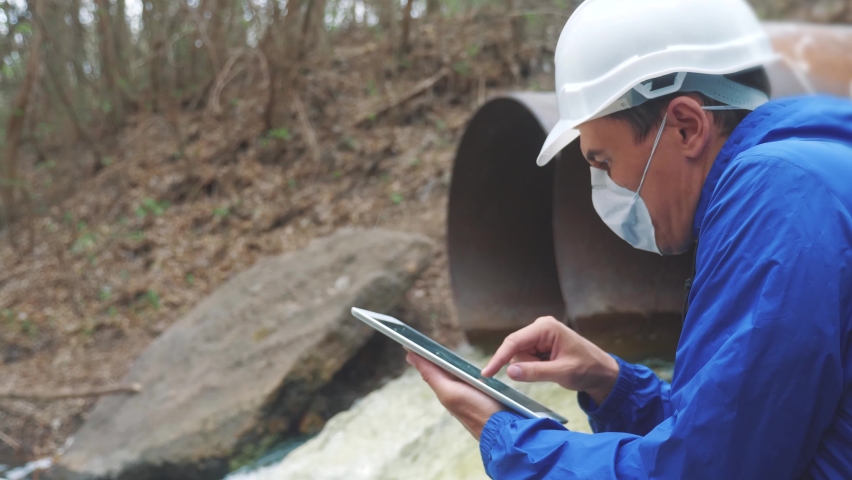 Environmental pollution radiation waste concept . man chemist scientist in a respirator mask studies water pollution digital tablet and an white helmet stands next to the waste pipes. conservation | Shutterstock HD Video #1058699596