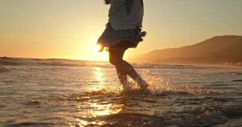 Slow motion woman feet walking barefoot by beach at golden sunset in the ocean waters. Female tourist on summer vacation in Malibu, California, USA. Woman in beautiful waving dress at sunset, 4K