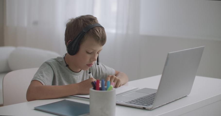 little boy is watching educational videos for children on laptop and listening by headphones, drawing on paper