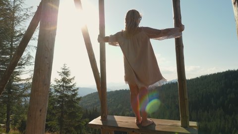Rear view of woman in dress swinging on large wooden swing with scenic mountain view at sunny summer day, slow-motion shot Stock Video