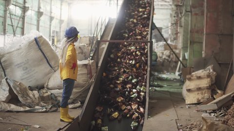 Woman-worker in yellow and transparent protecting glasses, hard hat and mask watching the conveyor full of used plastic bottles lifting up. Footage of automized process on recycle plant