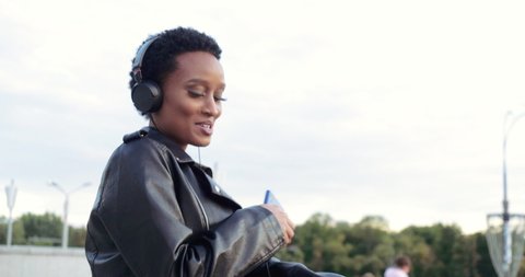 Active stylish African model wears headphones leather jacket dances walking down street to music, sings songs, moves her hand to melody, holds modern smartphone device, enjoys listening with earphones : vidéo de stock