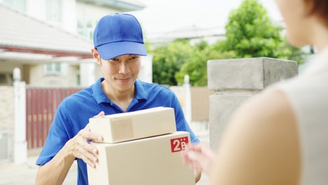 Young Asia postal delivery courier man in blue shirt handling parcel boxes for sending to customer at house and Asian female receive delivered package outdoors. Package shopping food delivery concept.