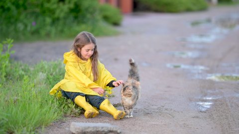 Happy Child in Yellow Raincoat and Boots Sits on the Grass and plays with the Cat.