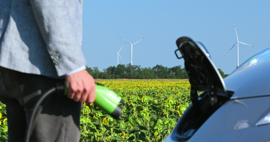 A man inserts a power cord into an environmentally friendly electric car, side close-up with focus to the background with wind turbines. Royalty-Free Stock Footage #1058719645