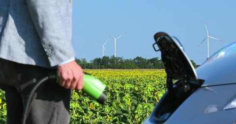 A man inserts a power cord into an environmentally friendly electric car, side close-up with focus to the background with wind turbines.