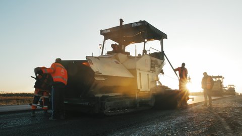 Novosibirsk region, September 4, 2020. Two road workers pour hot asphalt into a paver at sunset. Road construction machines. Road repair. Beautiful sun glare.
