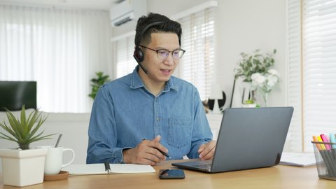 Smiling asian man wear wireless headset making conference videreo call on laptop. Male professional call center agent, hr manager having distance webcam chat job interview on computer at home office.