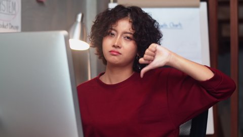 Unhappy Asian Businesswoman looking at camera and Doing Thumbs Down Hand Sign Gesture in creative office workplace. Female Worker Feels Disappointed Gesturing Dislike, Badly sign by using finger sign.