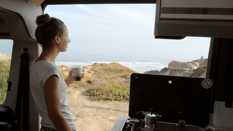 Young woman having a fresh cup of coffee in her camper van enjoys coastal views from her vehicle. Female solo traveller living in her van drinking coffee in the morning 