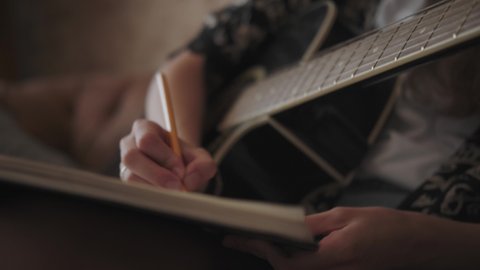 Creative female musician with guitar alone writing song, composing music sitting on sofa in bright day room. A woman plays the guitar, stops and writes text or notes in a notebook. To compose music