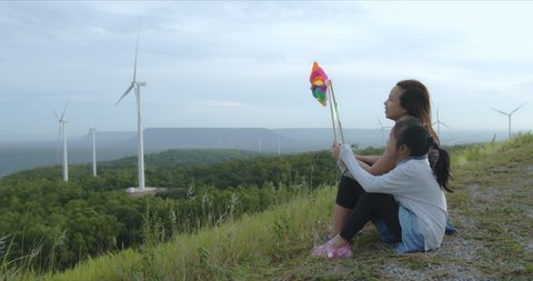 Asian mother and daughter are playing with turbine toy at the wind turbine field with fun together. Child girl and mother enjoy with beautiful scenery view of nature in slow motion shot.