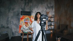 Female is showing paintbrushes and paints while recording tutorial video for her followers, standing against a portrait in concept art style