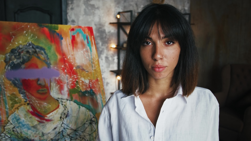 Female painter is holding paintbrushes and palette while posing by newly painted portrait in modern art style Royalty-Free Stock Footage #1058729185