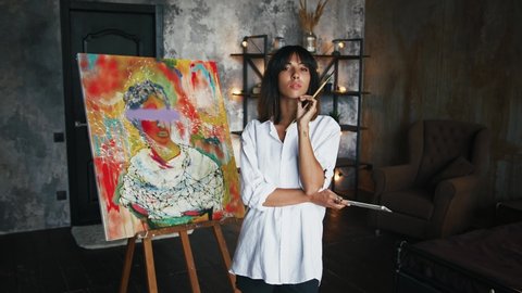 Female painter is holding paintbrushes and palette while posing by newly painted portrait in modern art style