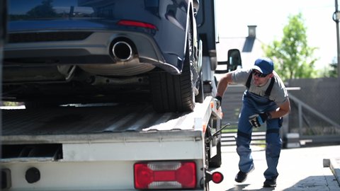 Tow Truck Male Driver Checking And Securing Car On Top Of Flatbed Towing Trailer. Car Transporter Ready To Depart.