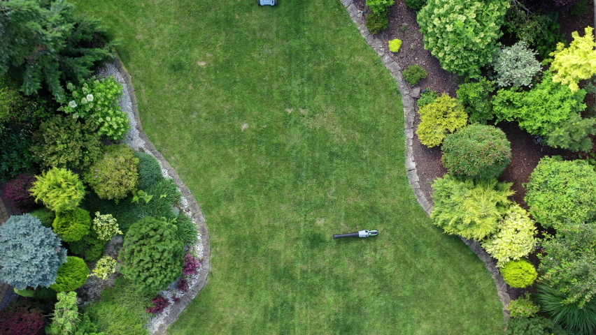 Large Green Grass Lawn Trees And Shrubs In Well Maintained Landscape And Garden. Maintenance Worker Cutting Grass With Mower.