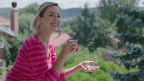 Blond haired young woman is drinking a hot tea while watching the green view on the balcony. Woman sniffs the tea and looks at the color of the tea. Slow motion video.