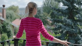 Young woman with blond hair is watching the green view on the balcony. Smiling, looking at the camera.Slow motion video.