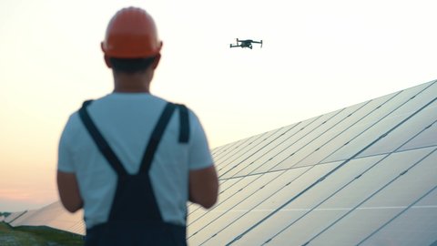 Industrial expert wearing helmet and controlling drone in photovoltaic solar power plant. Solar panel array installation. Technologies and ecology. Sunsets.