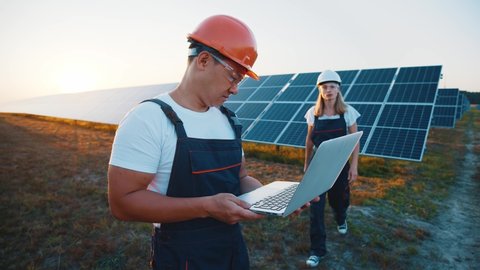 Asian developer engineer managing solar panel batteries by using portable laptop negotiating with female partner worker on industrial station. Solar farm. Green energy.