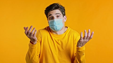 Young handsome unsure guy in sterile mask shrugs his arms, makes gesture of I don't know, can't help anything. Yellow studio background.